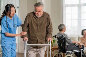 Older adult walking beside a handrail in a well-lit assisted living hallway with clear floors and supportive footwear.