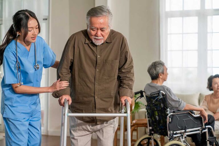 Older adult walking beside a handrail in a well-lit assisted living hallway with clear floors and supportive footwear.